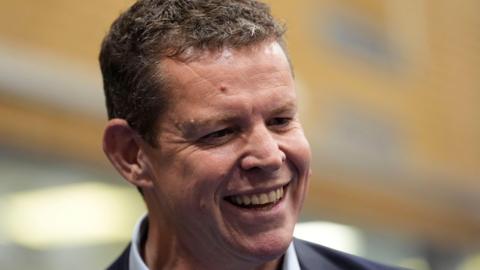A head and shoulders photograph of Rhun ap Iorwerth doing a media interview. He is smiling and looking to the right hand side of the picture. He has short brown hair and is wearing a light coloured shirt and dark suit.