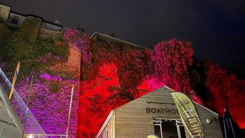The ivy-covered wall of Newquay Harbour is lit up with red, purple and pink lighting. In the foreground is a building with the letters BOATHOUSE on it and a flag advertising Cornish Rattler.