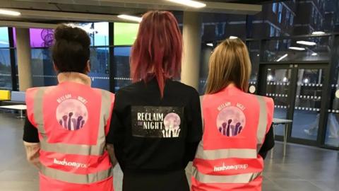 The backs of three women , two wearing high vis vests and one with a black top saying "Reclaim the Night"