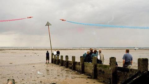 The Red Arrows flying above the water, which is at low tide, in Southend-on-Sea. One of the jets is blasting out a trail of red smoke, while the other has blue smoke. A small crowd of people are standing on mud watching them fly past.
