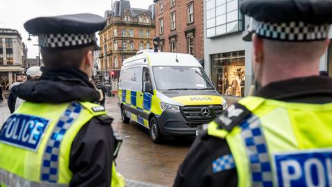 Two police officers stand with their backs to the camera, looking down Briggate in Leeds. They are wearing high-vis vests. They are looking at a police van, which is parked next to a shop.
