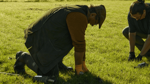 Two men are crouching down in a grassy field and planting wildflower seeds.
