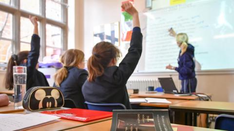 Three female students sitting in a classroom with their back to the camera while a female teacher writes on a board in the distance. The pupils are wearing school uniform and two have their right hands raised.