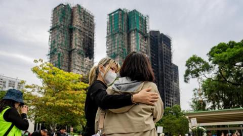A young woman in a dark jacket wearing a medical mask clasps another woman in a coat on a street on a cloudy day as charred apartment block towers loom in the distance. 