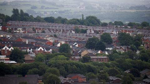 Rows of terraced houses and rooftops in Blackburn, there are a lot of trees to the front and the moors in the distance on an overcast day. 
