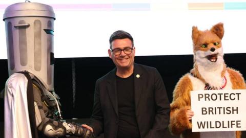 A smiling Andy Burnham stands alongside Count Binface and a costumed fox after being announced the winner of the Makerfield election.