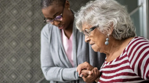 A woman helps and elderly woman through a house. She is holding her hand as she guides her. The older woman has a maroon and white stripey top on and earrings with grey hair. The carer has a pink shirt and grey cardigan on and glasses.