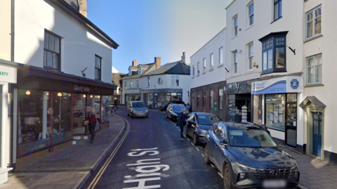 The high street in Sidmouth. There is a one way street with shops pictured either side of the road.