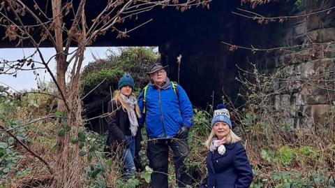Two blonde women and a man stand in overgrown greenery under a railway bridge