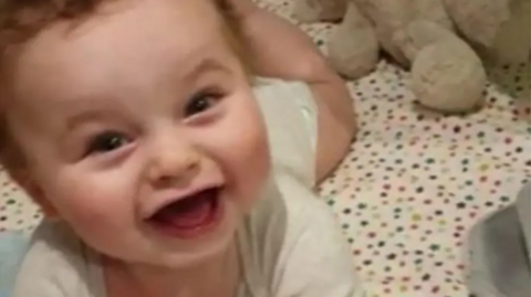 Baby Preston Davey smiles at the camera dressed in a white vest and appears to be lying in a cot with star bedsheet cover