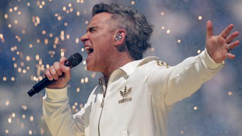 Robbie Williams performs in a white Adidas tracksuit with gold trim. He is holding a microphone and singing emphatically as pyrotechnics fall behind him.