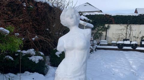 A classical Greek style sculpture of a woman with no arms made from snow. Behind the sculpture is a snowy suburban garden.