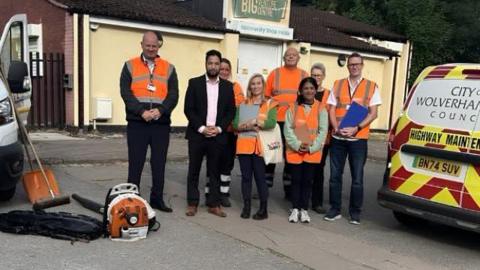 A group of people in high-vis vests stand next to a man in a suit, next to vans and outside a building