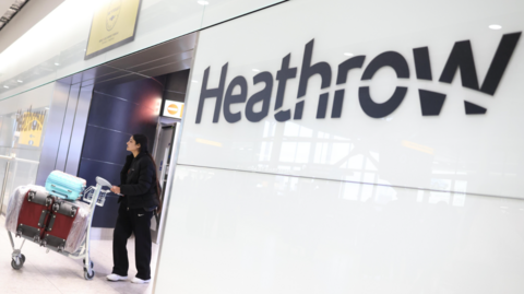 A woman walks through a doorway pushing a trolley with luggage stacked on it with the word Heathrow written in black beside her