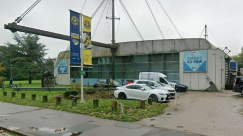 Google Street View of Oxford Ice Rink's exterior, a large white and blue building, with some moss growing on the roof. Large windows are at its centre.