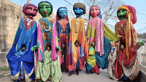 Seven towering, brightly coloured giant puppets in flowing patterned garments are carried by hidden performers along a narrow outdoor road, with a wall, fence, and bare tree in the background.