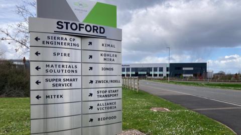A large metal sign listing some of the companies which are trading at Worcester Six business park. The sign is on a grassy verge with big buildings visible in the distance.