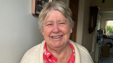 Menyw yn gweni at y camera ar ei stepen drws. Mae ganddi wallt gwyn ac yn gwisgo ffroc a cardigan gwyn. Yn y cefndir mae'n bosib gweld ei chyntedd a'i chegin.  