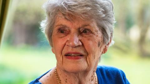 Jane Darlison, wearing a blue jumper and chain necklace, looking off-camera with a window to a garden behind her