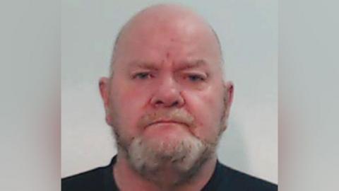 A man in his 50s with a bald head and a beard in a police mugshot