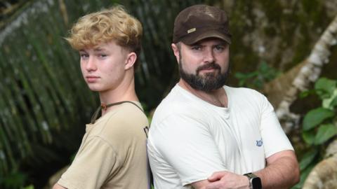 A boy with wavy blonde hair and a stone coloured t shirt stands back to back with a man in a white t shirt and brown cap