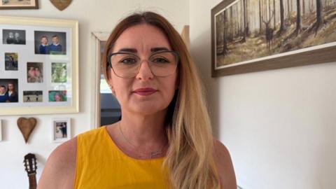 Rachel Townsend poses for the camera. she has blonde hair and is wearing a yellow top. she is stood in her phone with family pictures in the background
