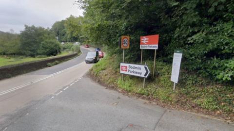 A road junction with two signs road saying Bodmin Parkway. The road has heavy foliage on one side and a field on the other, protected by a low granite wall. About half a dozen cars drive on one side of the road.