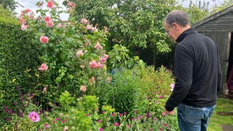 Man wearing glasses, black jumper and blue jeans in his garden overlooking a bed with colourful flowers.