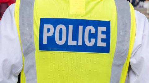 A generic shot of a police officer from behind. They are wearing a hi-viz vest with the word "police" printed on it. 