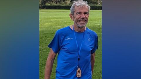 A man in a light blue running top smiles at the camera. He has grey hair and a grey beard. The top has parkrun written across his right chest in white. He is wearing a lanyard around his neck with a wooden pendant on the end, with 500 carved out of it.