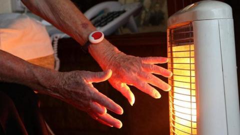 An elderly woman's hands outstretched towards a heater. Her hands are glowing yellow from the lamp.