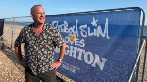 A man, Fatboy Slim, wearing a black and white floral shirt. He is stood in front of a beach, with a blue sign with white writing behind him saying Fatboy Slim loves Brighton.