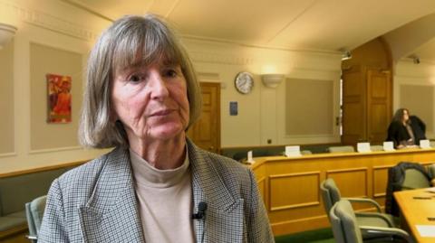 A woman stands inside a council chamber with a serious look on her face. She has grey hair and is wearing a check jacket. A large wooden bench surrounded by green chairs is visible in the background. The walls and ceiling are white with beige panels and a painting.