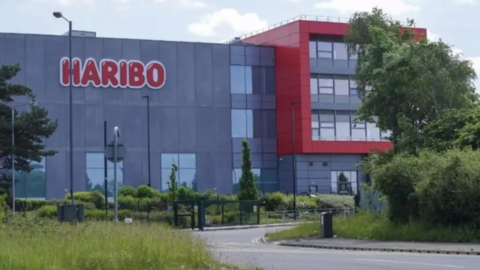 A modern industrial building with grey and red exterior panels displays a large “HARIBO” sign on the front. The site is surrounded by greenery, including grass, shrubs, and trees, with a road and gate leading up to the entrance.