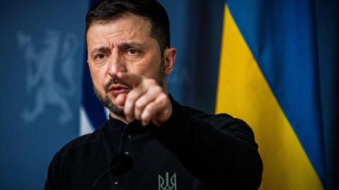 Volodymyr Zelensky speaking at a press conference in Oslo standing in front of a Ukrainian flag pointing.