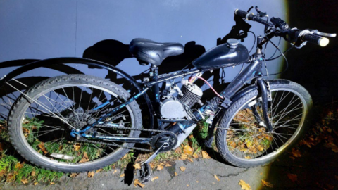 A bicycle is propped up against a wall at night. It is dark in colour and has an engine attached to the cross bar.