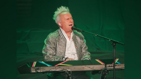 Howard Jones at a keyboard, performing. He is singing with his eyes closed. He has his trademark spiky blonde hair and a silver sparkled jacket.