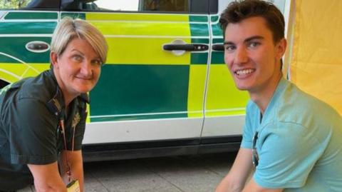 Claire Bird kneels beside her son James Bird in front of a green and yellow ambulance vehicle. Both are smiling at the camera.