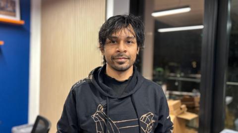 Man wearing a dark grey hoodie looks at the camera. He has black hair, slight brushed forward fringe and a short beard growth. He is in what looks like an office, with a window looking out to a balcony.