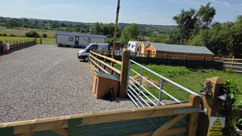 An open area of land with a gravel surface. There is a mobile home on the site along with a caravan and two sheds. In the background are hills and greenery.