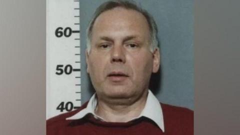 A police mugshot of a man with short greying hair, wearing a shite shirt and a red jumper.
