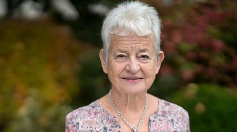 Jacqueline Wilson, best-selling children's author, at the Cheltenham Literature Festival. Jacqueline has grey hair and is wearing a silver chain and pink and blue shirt with flowers.