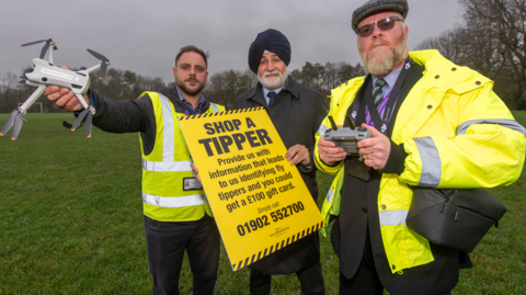 Thomas Hawkins, section leader in fly tipping, councillor Bhupinder Gakhal, cabinet member for resident services and Ian Crutchley, senior compliance officer. The men are stood in a field holding the drone and a council posted about reporting fly tipping.