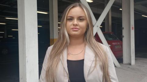 A woman with long brown hair stands in front of a car park