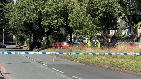 Police tape across a road and behind it was can see a smashed up car against a tree with two people in high-vis jackets next to it. The car is surrounded by several trees