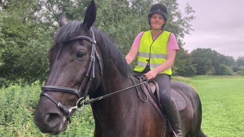 A female horse rider, wearing a pink t-shirt, black jodhpurs, a black riding hat and yellow hi-viz, is sitting on a dark bay horse in a field alongside a row of trees.  
