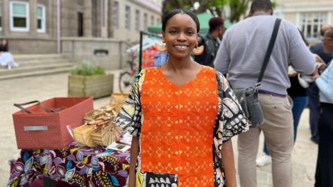 Seun smiles at the camera next to a stall with a traditional African cover over it with baskets on top of it