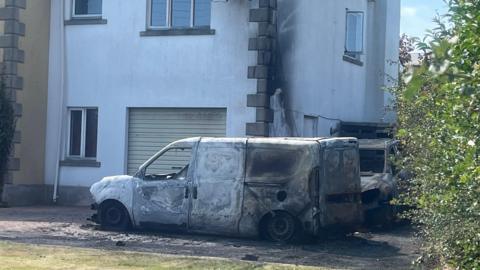 A white van in front of a house. Both the van and the house show sings of fire damage. There is a car behind the van, which also displays signs of fire damage. 