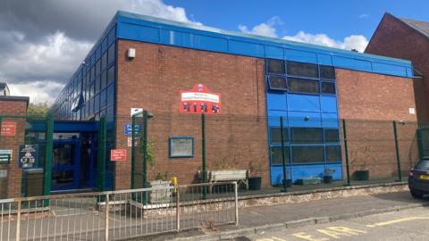 A red brick block building with blue windows behind a green fence 