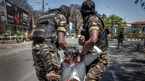 Two uniformed members of Madagascar's security forces detain a protester, who wears a surgical mask and a red whistle around his neck
